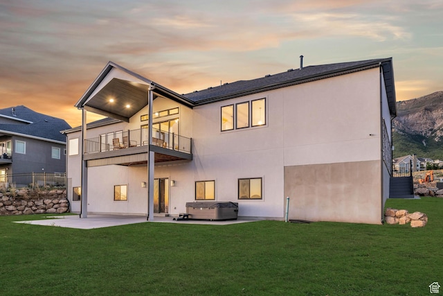 back of property at dusk featuring stucco siding, a hot tub, a yard, a balcony, and a patio area