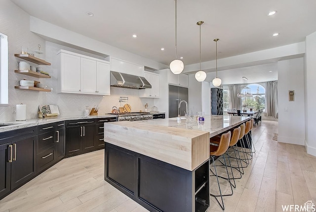 kitchen with wall chimney exhaust hood, tasteful backsplash, white cabinetry, open shelves, and a breakfast bar area