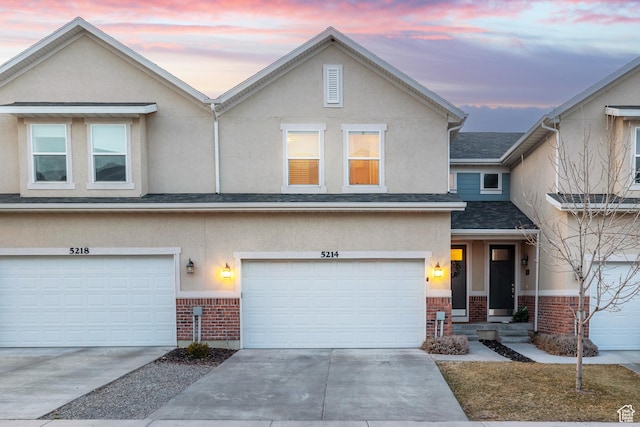 multi unit property with driveway, stucco siding, brick siding, a shingled roof, and a garage