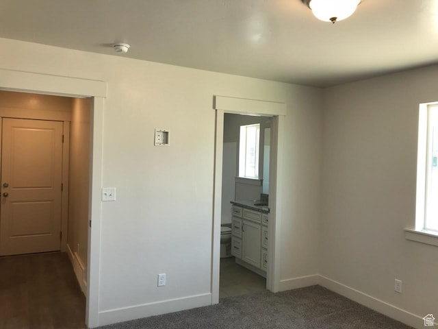unfurnished bedroom with multiple windows, connected bathroom, baseboards, and dark colored carpet