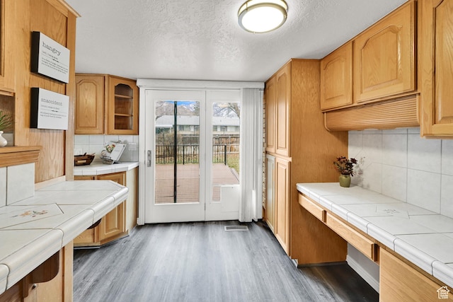 kitchen with tile counters, decorative backsplash, a textured ceiling, glass insert cabinets, and wood finished floors