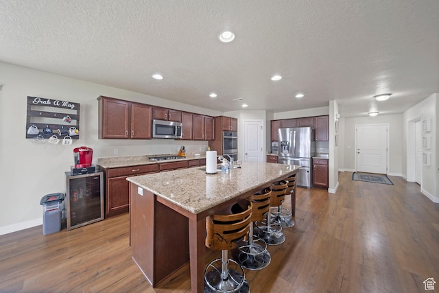 kitchen with beverage cooler, a breakfast bar, a kitchen island with sink, stainless steel appliances, and dark wood-type flooring