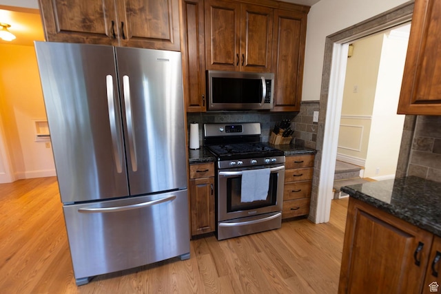 kitchen with backsplash, appliances with stainless steel finishes, dark stone counters, and light wood-style floors