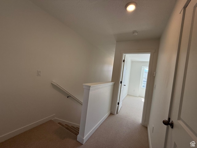 corridor featuring baseboards, an upstairs landing, light colored carpet, and a textured ceiling