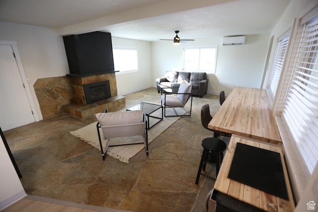 living room with a ceiling fan, a wall unit AC, a stone fireplace, stone finish floor, and baseboards