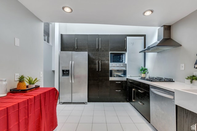 kitchen with light countertops, wall chimney exhaust hood, dark cabinetry, appliances with stainless steel finishes, and light tile patterned floors