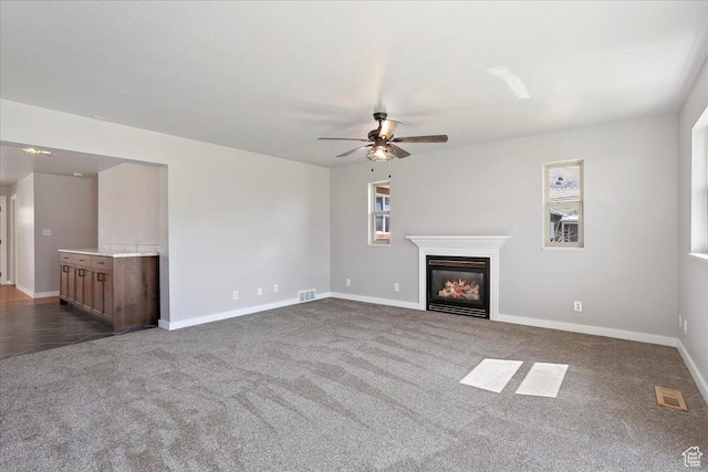 unfurnished living room featuring visible vents, ceiling fan, dark carpet, and a glass covered fireplace