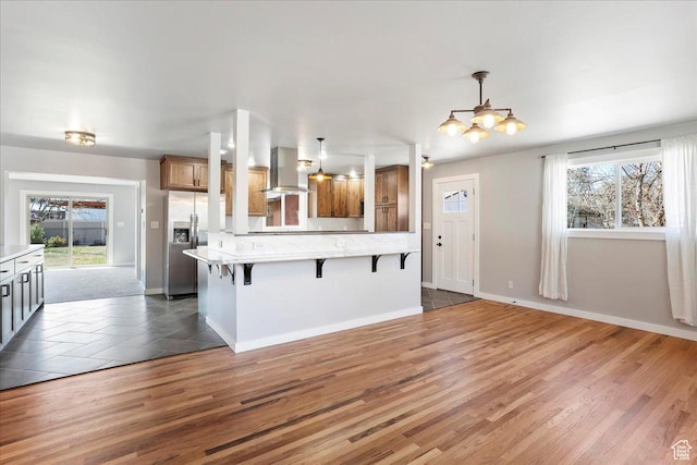 kitchen with stainless steel fridge with ice dispenser, a breakfast bar area, wall chimney exhaust hood, wood finished floors, and brown cabinetry