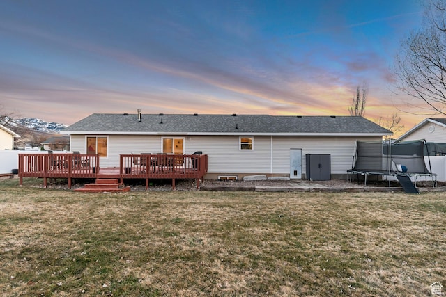 back of house at dusk with a lawn, a trampoline, a wooden deck, and roof with shingles