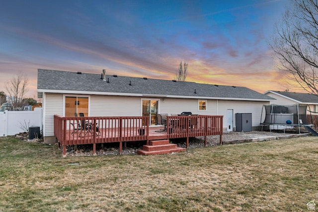 rear view of house featuring a trampoline, a deck, fence, and a lawn