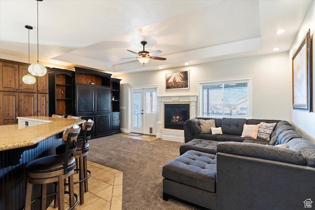living area featuring light colored carpet, a tiled fireplace, a ceiling fan, and a raised ceiling