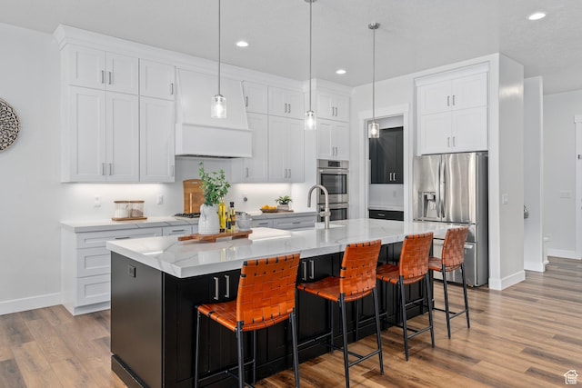 kitchen featuring white cabinets, appliances with stainless steel finishes, light wood-style flooring, a kitchen island with sink, and decorative light fixtures