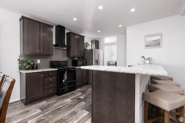 kitchen featuring a breakfast bar, wall chimney range hood, a peninsula, dark brown cabinetry, and black appliances