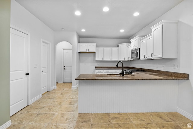 kitchen with white cabinets, a peninsula, stainless steel microwave, dark stone countertops, and a sink