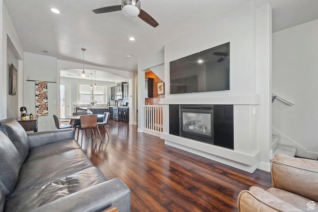living area featuring stairs, a glass covered fireplace, ceiling fan, recessed lighting, and dark wood-style flooring