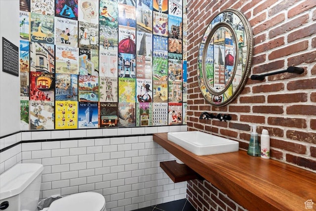 bathroom with a sink, tile walls, brick wall, a wainscoted wall, and toilet