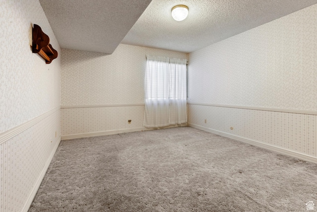 unfurnished room featuring a textured ceiling, wainscoting, baseboards, wallpapered walls, and carpet