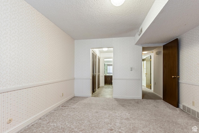 carpeted empty room featuring visible vents, a textured ceiling, wallpapered walls, and wainscoting