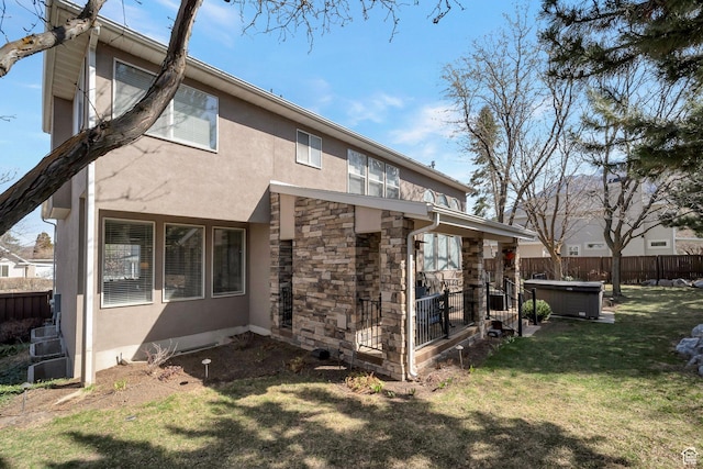 rear view of house with stucco siding, fence, a hot tub, stone siding, and a lawn