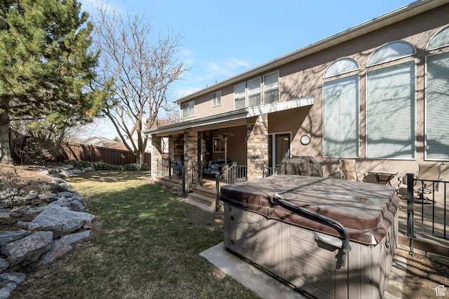 back of property featuring stone siding, fence, stucco siding, a hot tub, and a yard