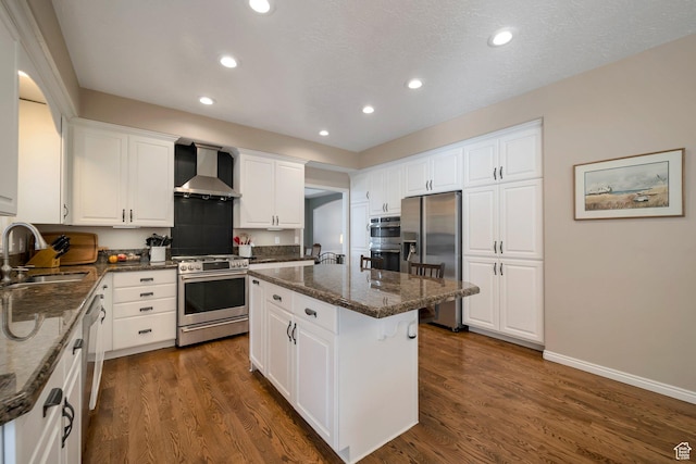 kitchen with dark wood finished floors, a kitchen island, a sink, appliances with stainless steel finishes, and wall chimney exhaust hood
