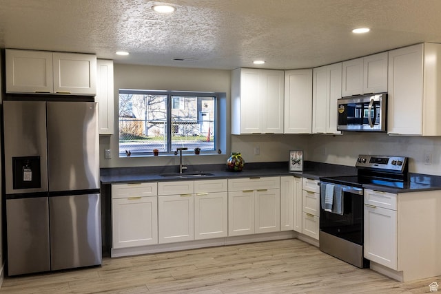 kitchen with dark countertops, appliances with stainless steel finishes, white cabinetry, a sink, and light wood-style floors