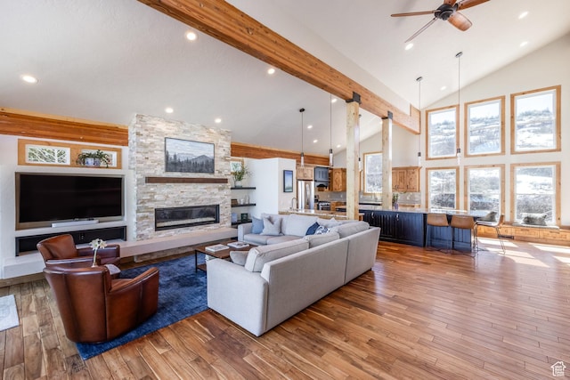 living area featuring ceiling fan, a stone fireplace, hardwood / wood-style flooring, high vaulted ceiling, and beamed ceiling
