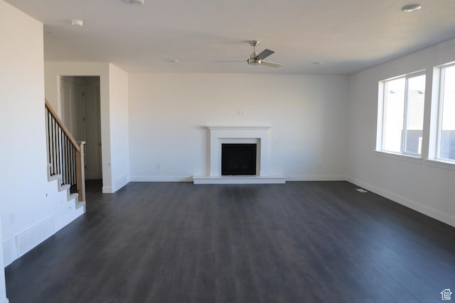 unfurnished living room with stairs, a ceiling fan, a fireplace with raised hearth, dark wood finished floors, and visible vents