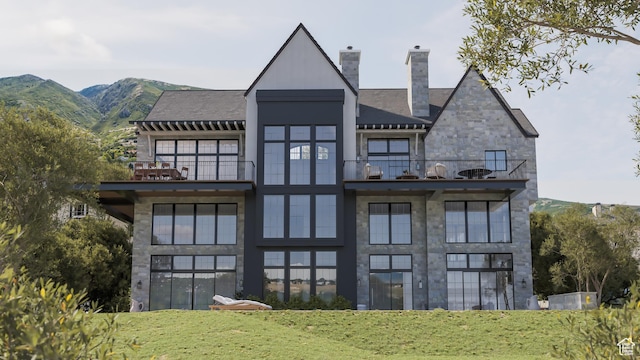 back of property with a balcony, stone siding, a chimney, a mountain view, and a yard