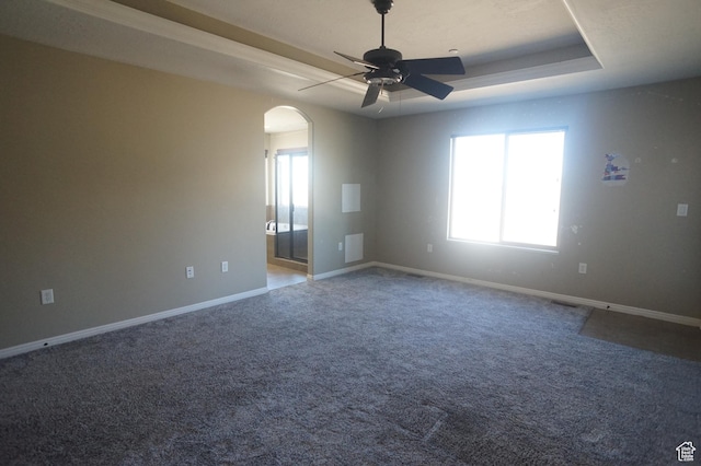 carpeted spare room featuring a tray ceiling, baseboards, arched walkways, and a ceiling fan