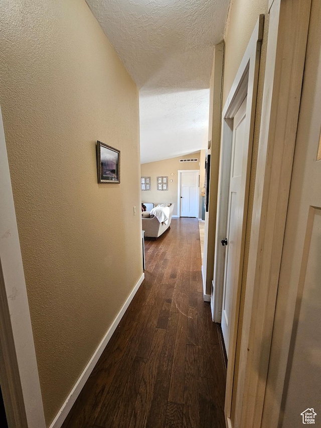 hall with vaulted ceiling, a textured wall, baseboards, a textured ceiling, and dark wood finished floors