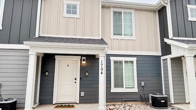view of exterior entry with a shingled roof, central AC, and board and batten siding