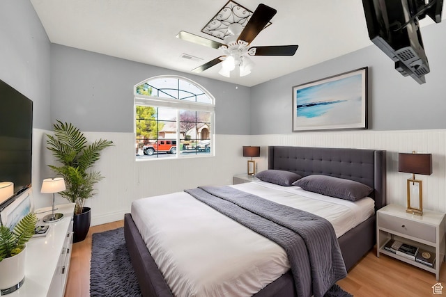 bedroom with a wainscoted wall, ceiling fan, visible vents, and light wood finished floors