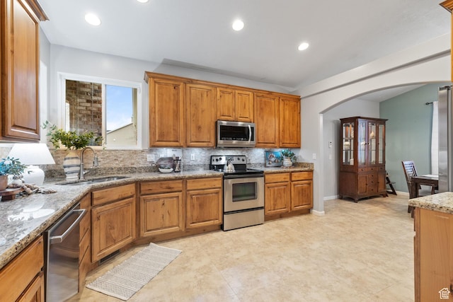 13968 S Hawberry Rd E, Draper, UT 84020 - kitchen featuring a sink, arched walkways, light stone countertops, appliances with stainless steel finishes, and brown cabinetry