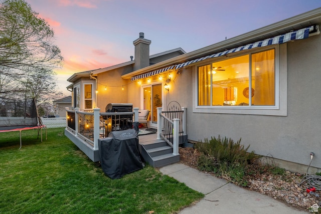 13968 S Hawberry Rd E, Draper, UT 84020 - back of house at dusk with a wooden deck, a chimney, a trampoline, stucco siding, and a lawn