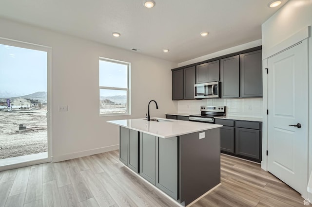 kitchen with light wood-style flooring, backsplash, a center island with sink, appliances with stainless steel finishes, and a sink