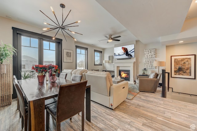 dining area featuring light wood finished floors, ceiling fan with notable chandelier, and a glass covered fireplace