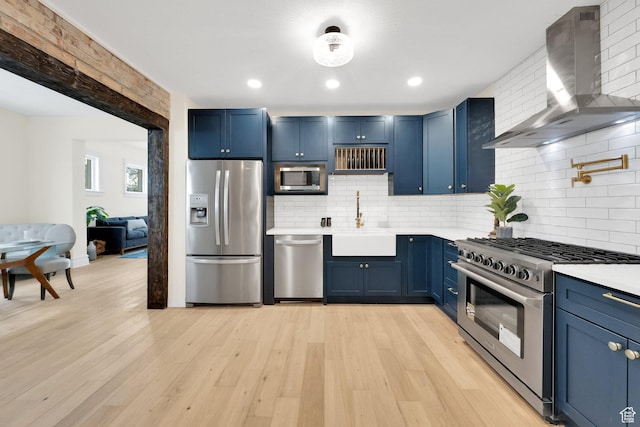 kitchen with blue cabinetry, light countertops, a sink, wall chimney range hood, and appliances with stainless steel finishes