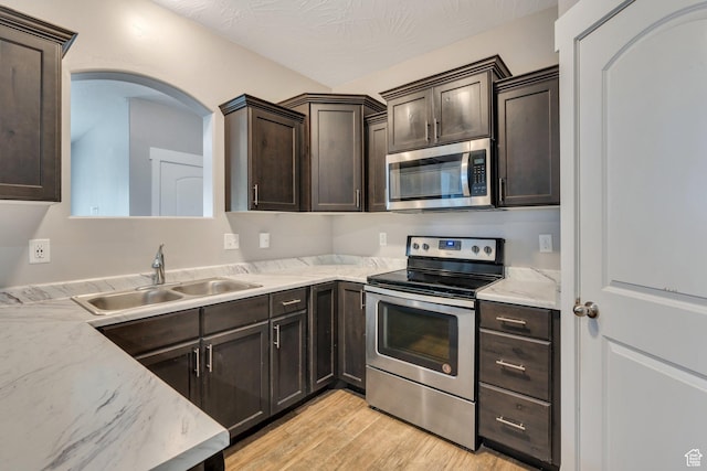 kitchen featuring appliances with stainless steel finishes, a sink, dark brown cabinets, light wood finished floors, and arched walkways
