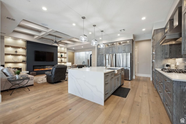 kitchen with stainless steel gas stovetop, light wood-style flooring, a ceiling fan, wall chimney range hood, and coffered ceiling