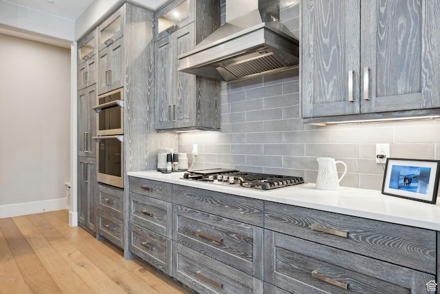 kitchen with light wood-style flooring, stainless steel appliances, wall chimney exhaust hood, tasteful backsplash, and gray cabinetry