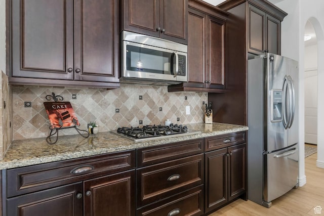 kitchen with dark brown cabinets, tasteful backsplash, appliances with stainless steel finishes, light wood-type flooring, and light stone counters