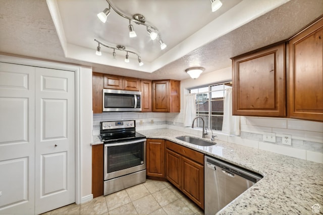 kitchen featuring decorative backsplash, appliances with stainless steel finishes, a sink, a tray ceiling, and brown cabinetry