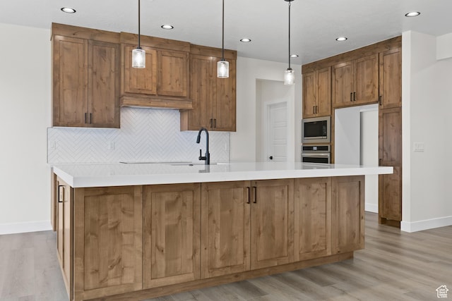 kitchen featuring a center island with sink, brown cabinets, light wood-type flooring, stainless steel appliances, and decorative backsplash