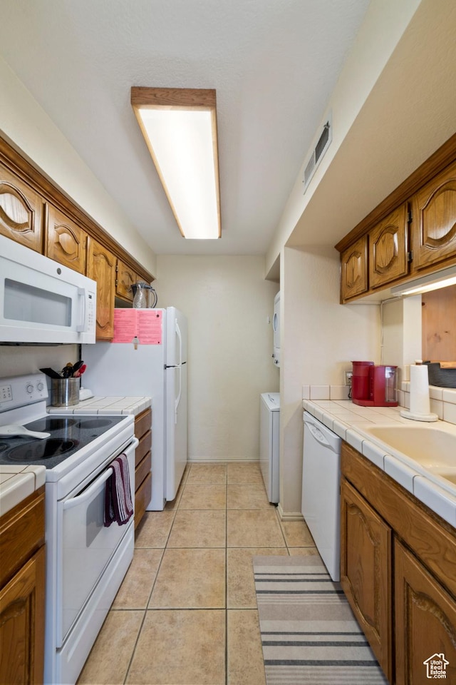 kitchen with tile counters, white appliances, light tile patterned floors, stacked washer and dryer, and brown cabinetry