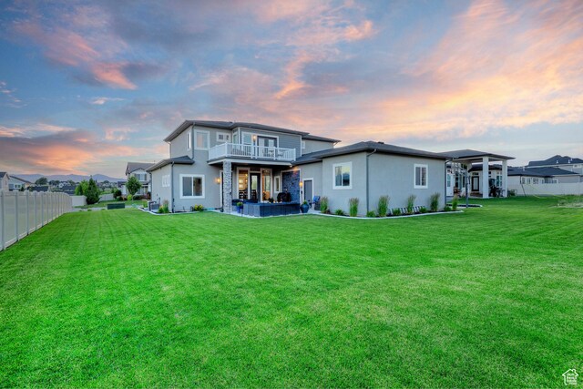 back of house at dusk featuring a balcony, fence, a patio, stucco siding, and a lawn