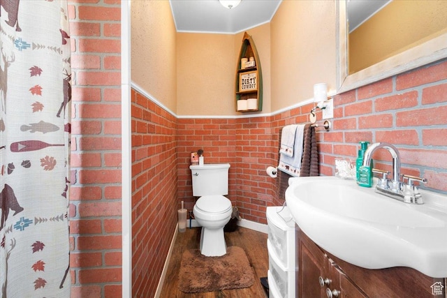 bathroom with toilet, vanity, wood finished floors, wainscoting, and brick wall