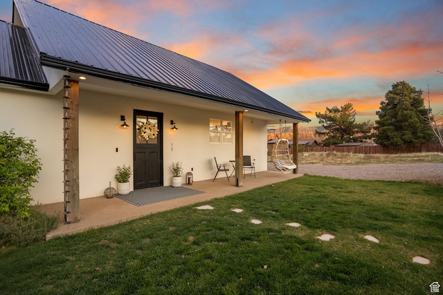 back of house at dusk with metal roof, a lawn, fence, a patio, and stucco siding