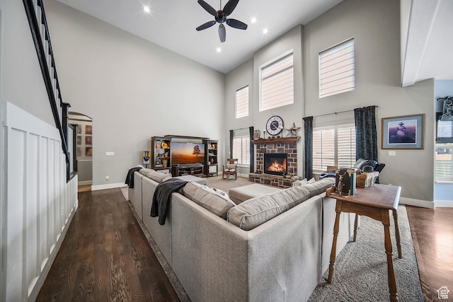 living area with ceiling fan, a towering ceiling, a stone fireplace, dark wood-style flooring, and baseboards
