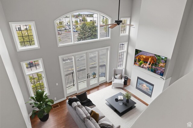 living area with a ceiling fan, a glass covered fireplace, a high ceiling, wood finished floors, and a healthy amount of sunlight